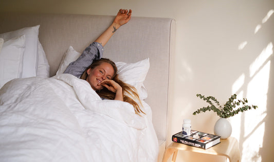 Woman smiling and stretching in bed in soft morning light beside a nightstand with a supplement bottle and vase of eucalyptus