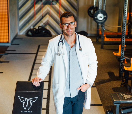Dr. Greene smiling in a white coat with stethoscope, leaning on a Monarch workout bench in a gym setting