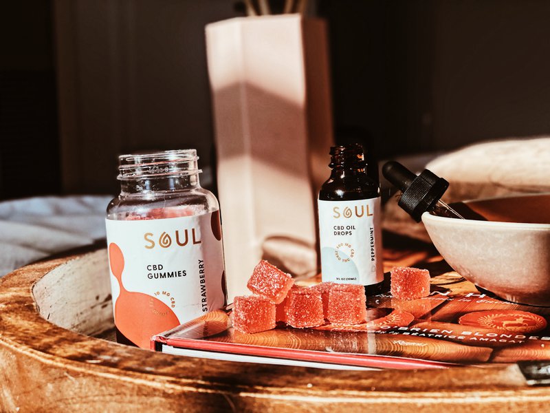 Close-up of Soul CBD gummies jar (strawberry) and Soul CBD oil dropper bottle with gummies arranged on a wooden tray in warm bedside light