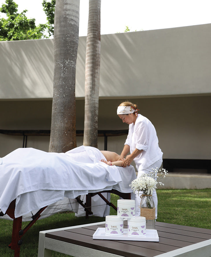 Massage therapist applying topical cream to a client’s leg on an outdoor massage table with Soul CBD jars and flowers nearby