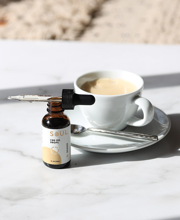 CBD oil dropper bottle beside a white coffee cup and saucer on a marble countertop in natural sunlight