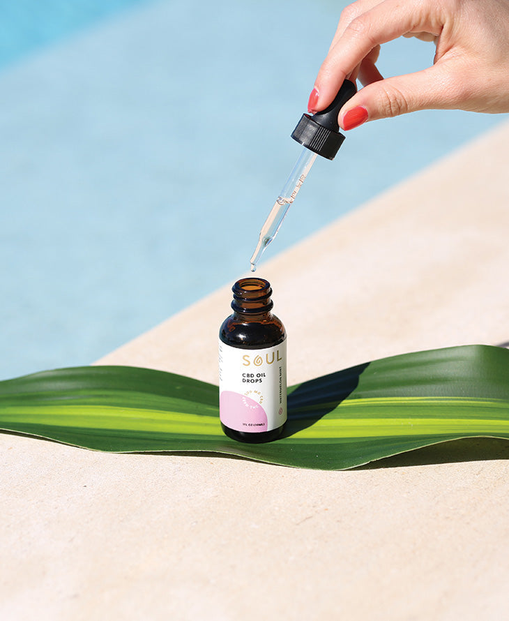 Hand holding a dropper releasing a drop above a Soul CBD Oil Drops bottle resting on a green leaf beside a pool
