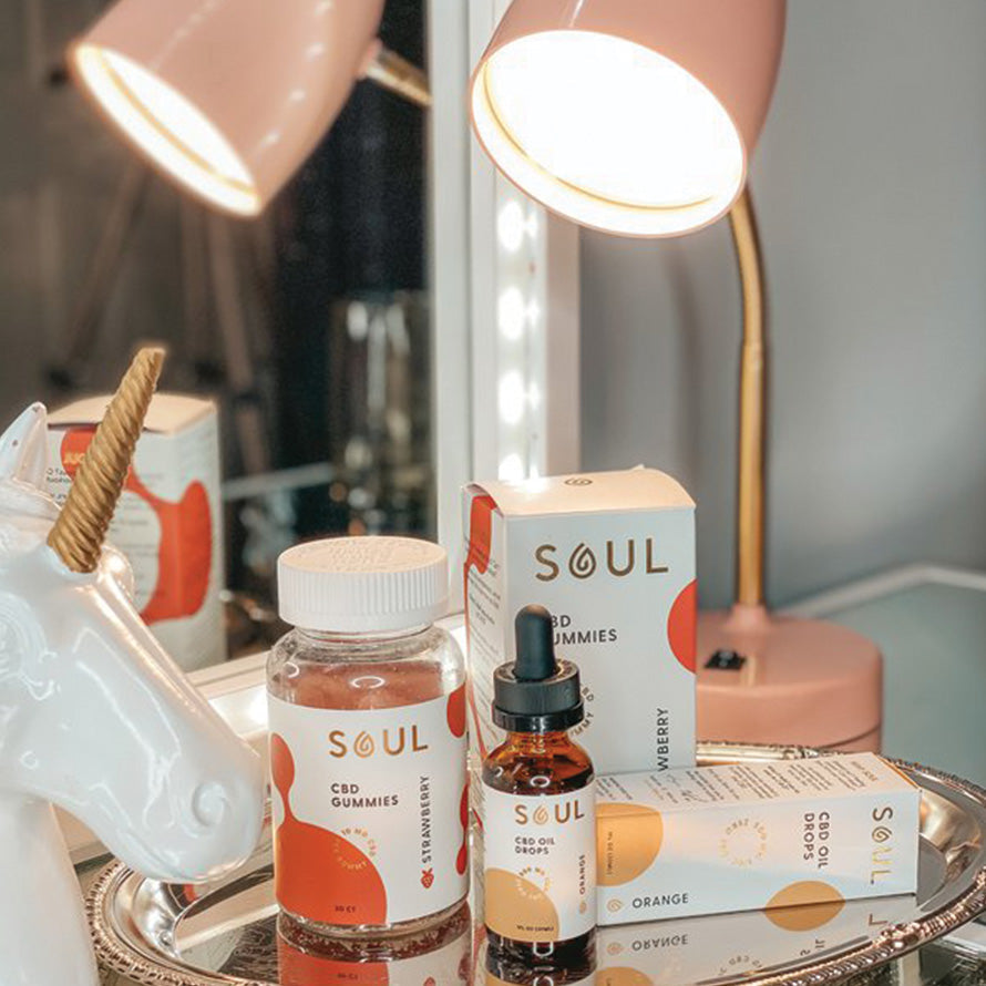 Soul CBD gummies jar and CBD oil dropper bottle displayed on a mirrored vanity tray beside a pink lamp and ceramic unicorn figurine