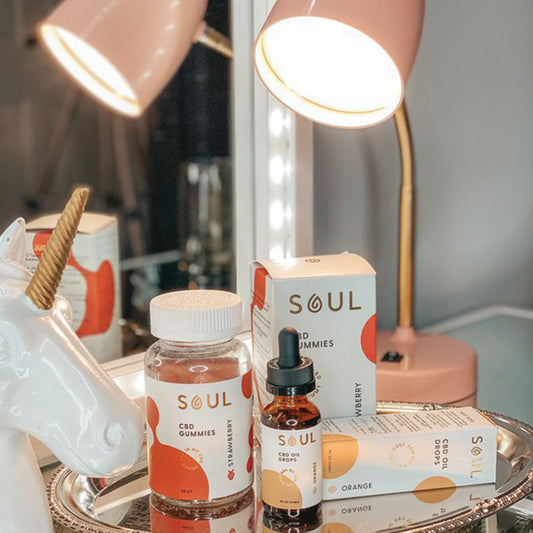 Soul CBD gummies jar and CBD oil dropper bottle displayed on a mirrored vanity tray beside a pink lamp and ceramic unicorn figurine