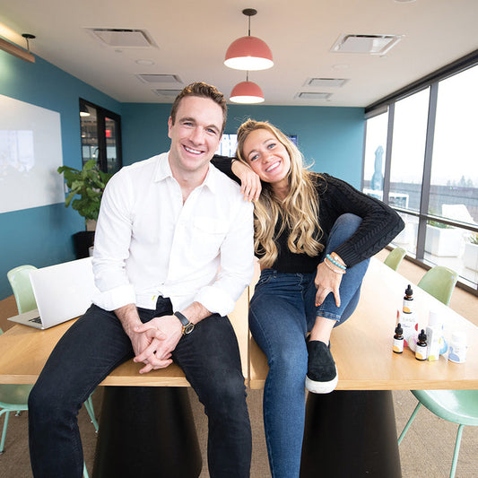 Two Soul CBD founders sitting on a conference table smiling with Soul CBD bottles and a laptop in a bright office with floor-to-ceiling windows