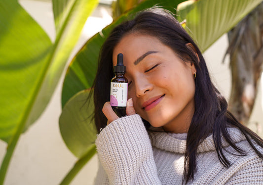 Woman holding a bottle of Soul CBD oil drops to her cheek, eyes closed, outdoors with large green leaves in the background