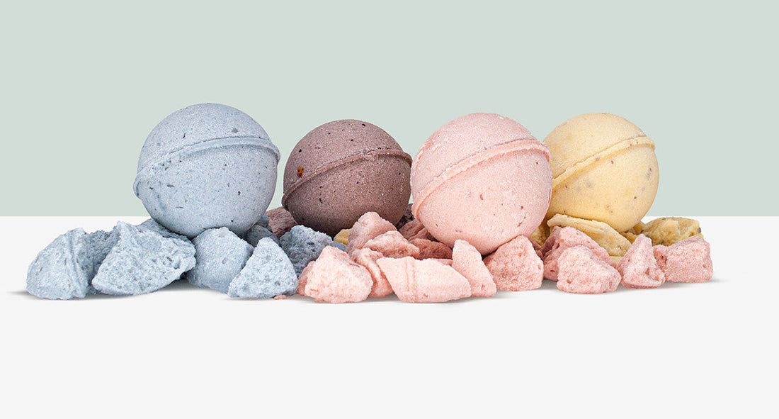 Front view of four CBD bath bombs in blue, purple, pink, and yellow resting on crumbled pieces against a pale mint background