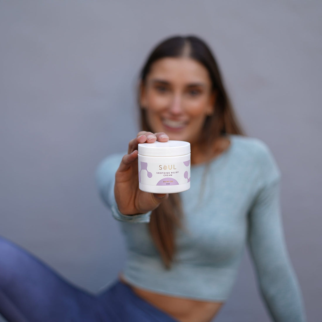 Woman holding a jar of SOUL Soothing Relief Cream toward the camera with a blurred gray background
