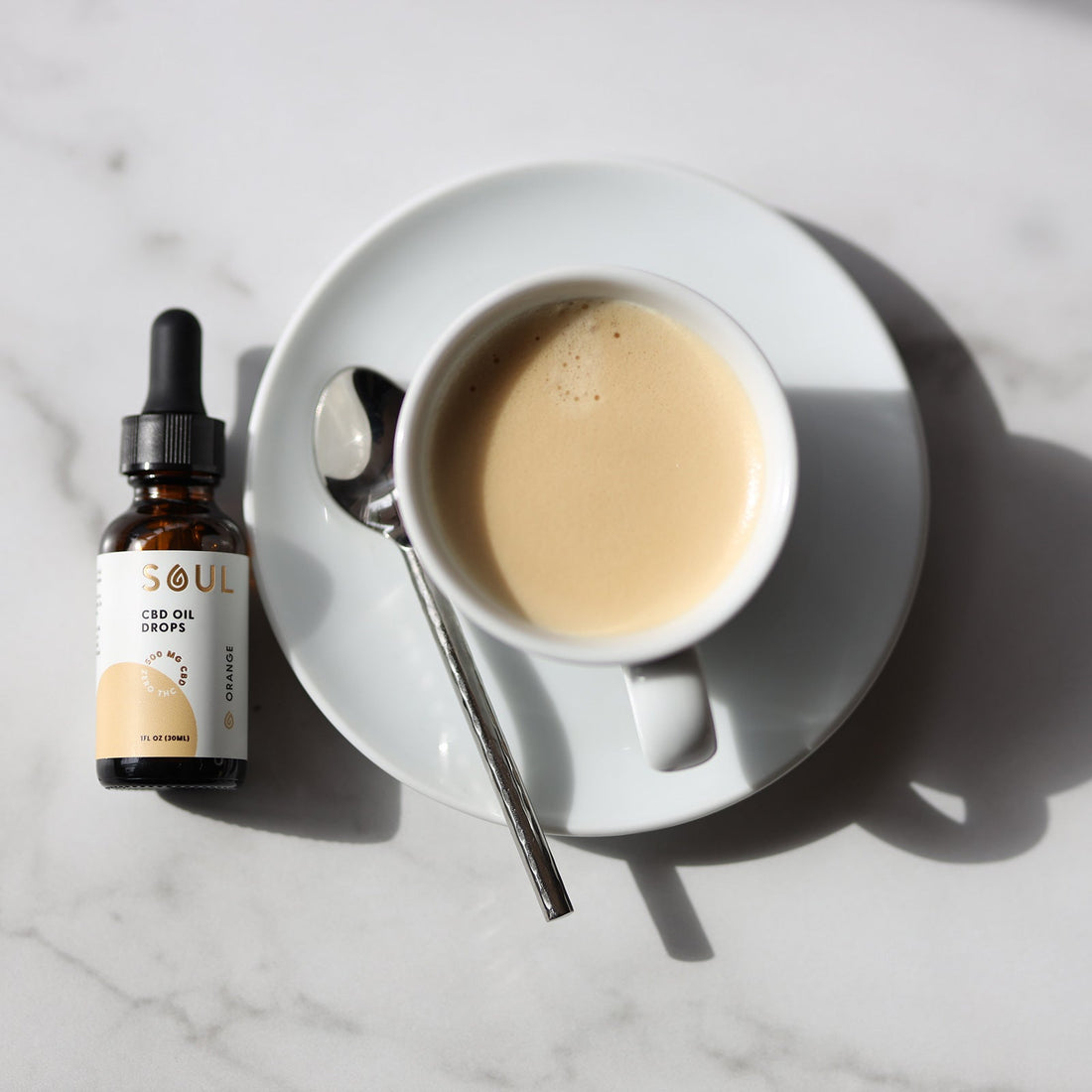 Top-down view of a cup of coffee with a spoon paired with a CBD oil dropper bottle on a marble tabletop
