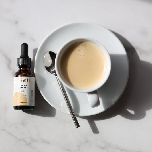 Top-down view of a cup of coffee with a spoon paired with a CBD oil dropper bottle on a marble tabletop