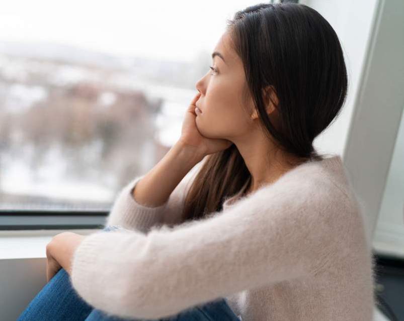Young woman sitting by a window looking pensive, gazing at a snowy cityscape outside