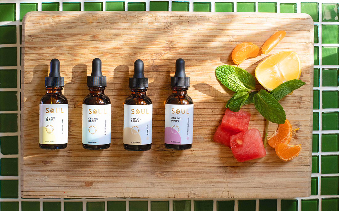 Four Soul CBD oil bottles on a wooden board beside mint, lemon, mandarin and watermelon over green tile