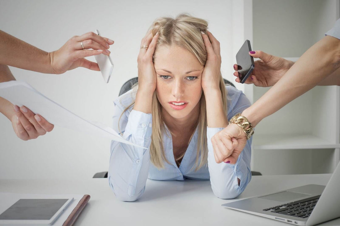 Woman at a desk holding her head, looking overwhelmed as hands offer phones, papers, and a watch to show work stress