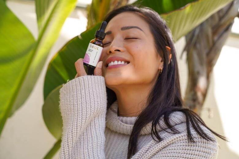 Smiling woman holding a Soul CBD tincture bottle to her cheek outdoors with large tropical leaves in the background