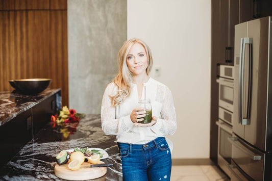 Woman holding a jar of green juice in a modern kitchen, standing by a marble countertop with cut lemons, an avocado, and a salad