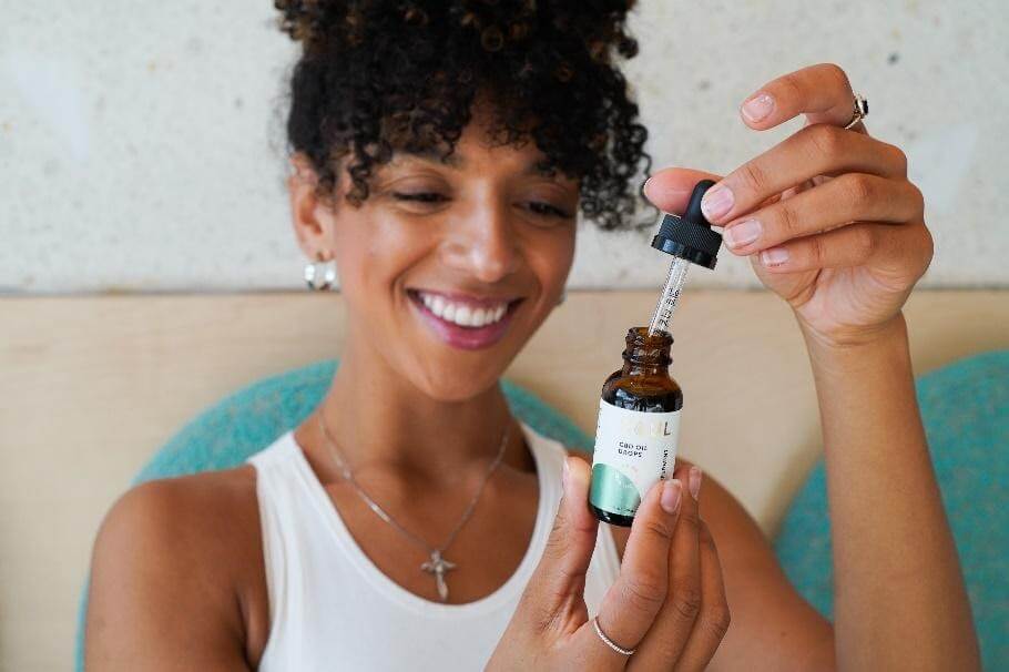 Smiling woman holding a CBD oil dropper over an amber bottle in a bright indoor setting