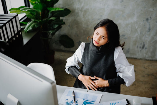 Woman at an office desk clutching her stomach in discomfort, suggesting bloating, with a computer, papers, and plant nearby