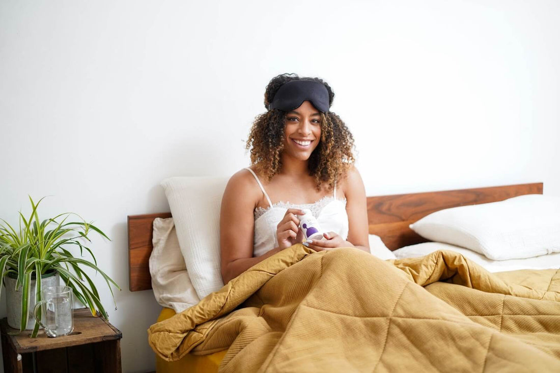 Smiling woman sitting up in bed with a sleep mask pushed to her forehead holding a bottle of supplements in a bright, minimal bedroom