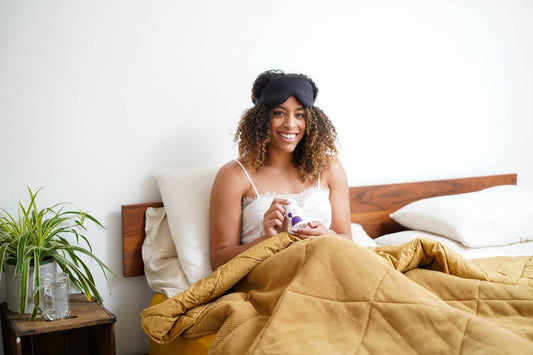 Smiling woman sitting up in bed with a sleep mask pushed to her forehead holding a bottle of supplements in a bright, minimal bedroom