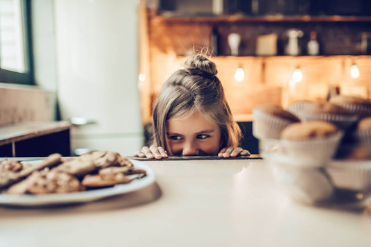 Young girl peeking over a countertop at a plate of cookies with warm kitchen lights in the background