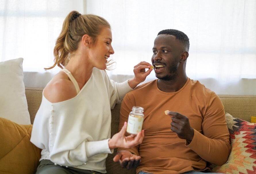 Woman offering a CBD gummy from a jar to a smiling man seated on a couch in a bright living room
