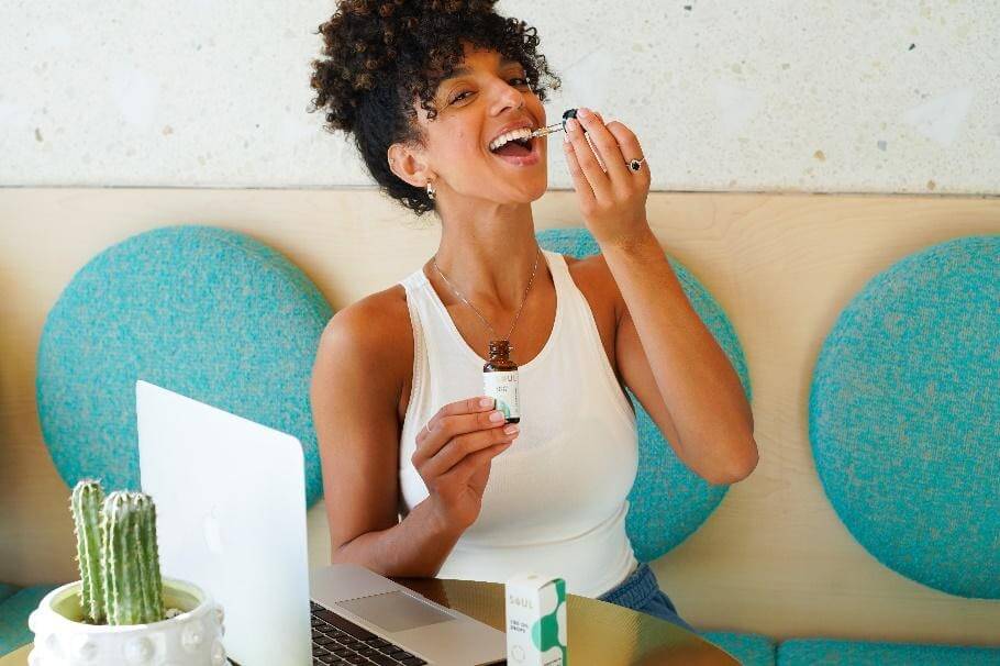 Woman using a CBD oil dropper while holding the bottle at a table with a laptop and product box, smiling