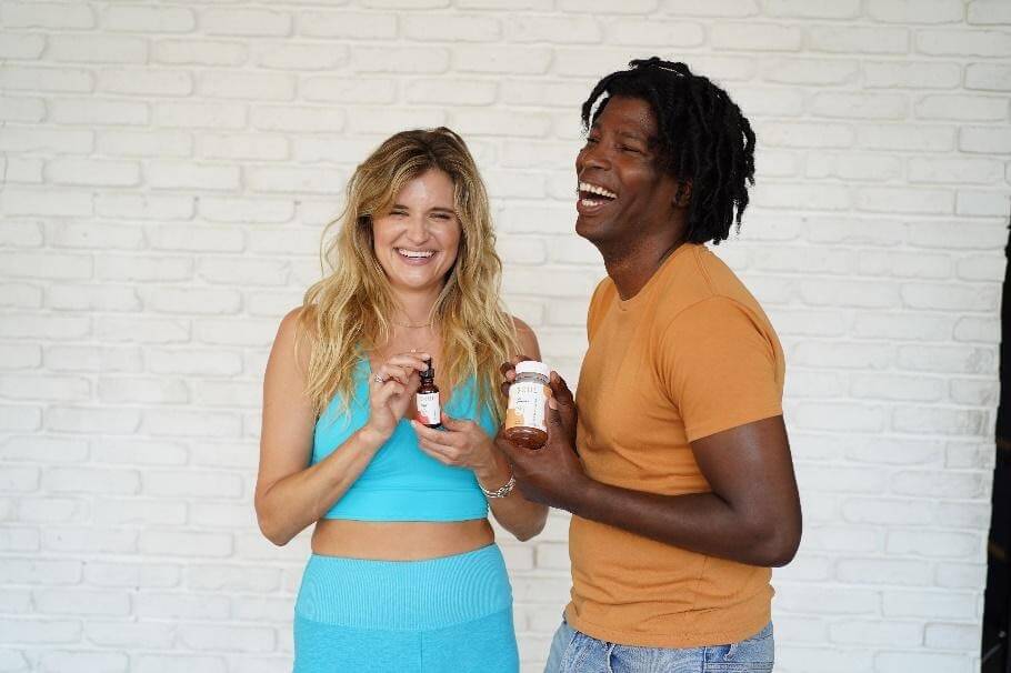 Woman in turquoise set smiles while holding a CBD oil dropper; man in orange t-shirt laughs holding a supplement jar against white brick wall