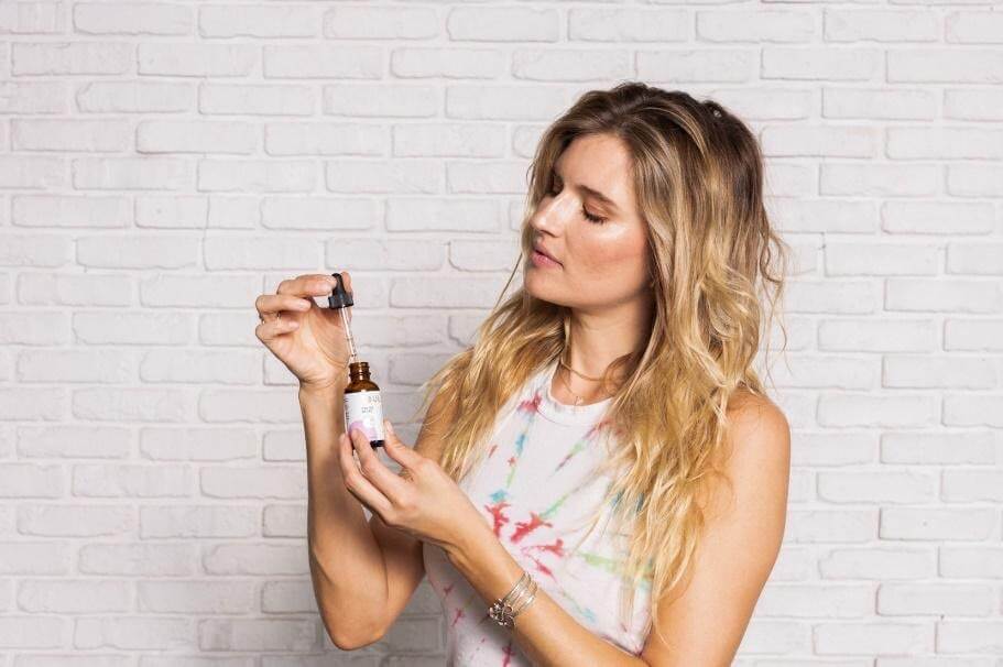 Woman inspecting a CBD oil dropper and amber bottle against a white brick wall background
