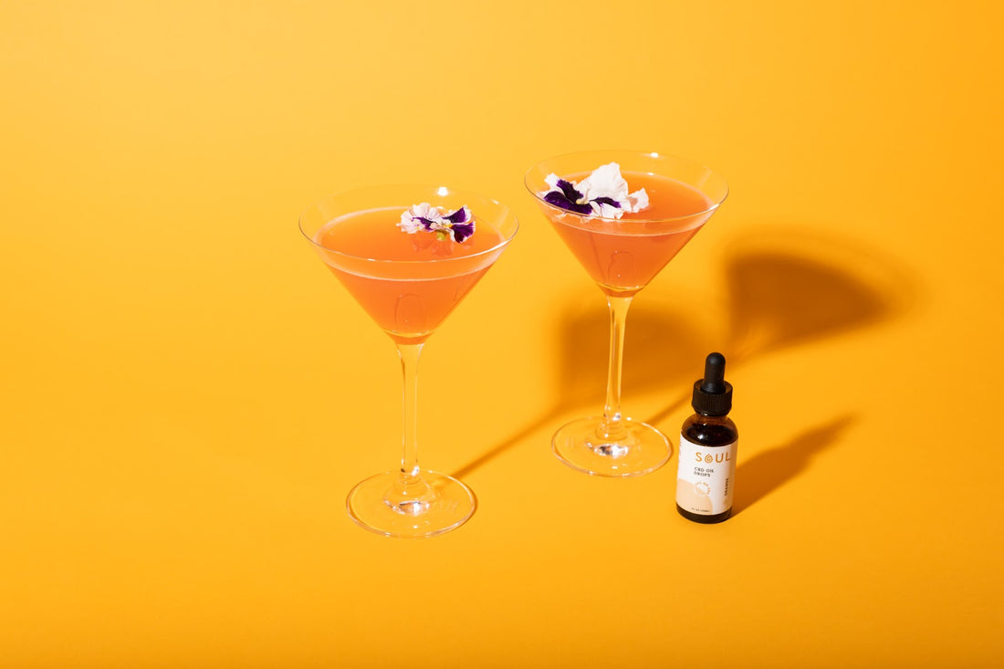 Two orange-hued CBD cocktails garnished with purple-and-white edible flowers beside a small Soul CBD oil dropper bottle on a bright yellow background
