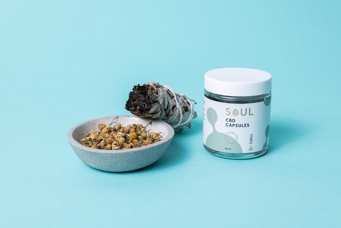 Front view of Soul CBD Capsules jar labeled CBD Capsules Chill beside a bowl of dried chamomile and a sage smudge on a light blue background