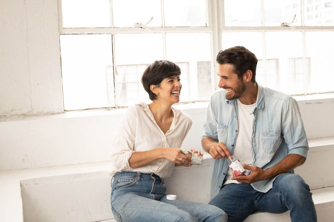 Two people laughing and sharing CBD supplement jars while seated by a bright window