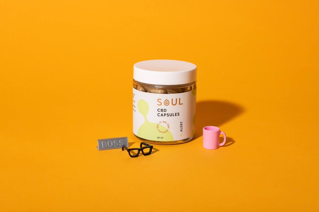 Front view of Soul CBD Capsules jar showing label, white lid, visible capsules and “30 CT” marking with mini pink mug and glasses on orange background