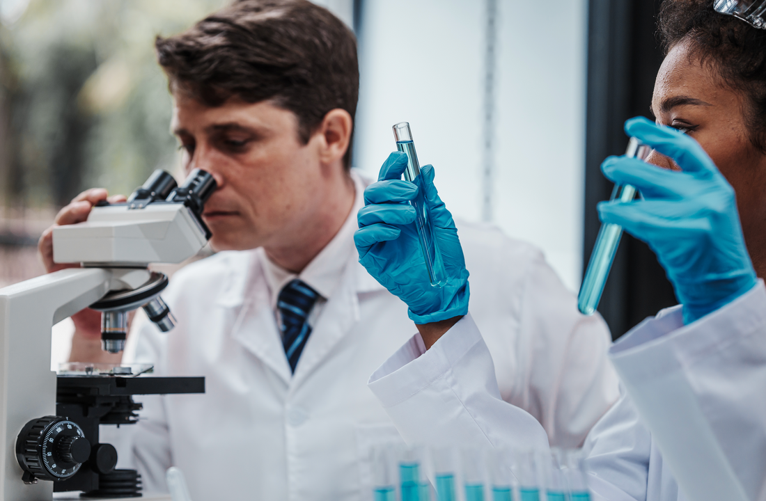 Two laboratory scientists examining blue liquid in test tubes while one looks through a microscope in a lab setting