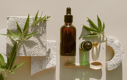 Amber tincture bottle with dropper, cannabis leaves, and a wooden spoon on textured white stone under soft natural light