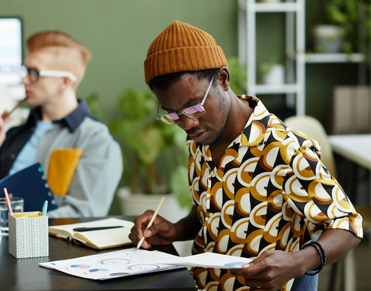 Man in a mustard beanie and patterned shirt reviewing charts and taking notes at a desk in a modern office