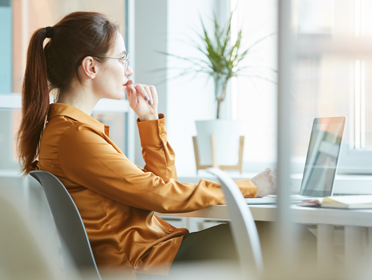 Woman in a mustard blouse sitting at a desk, looking thoughtfully at a laptop in a bright office