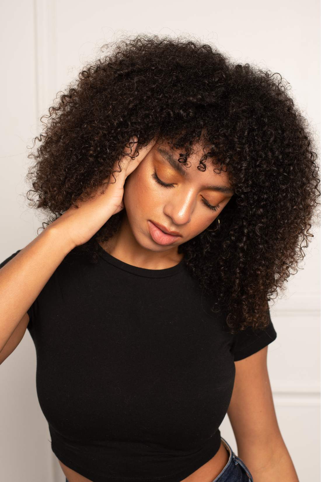 Woman with curly hair touching her temple and closing her eyes, appearing stressed in a neutral indoor portrait