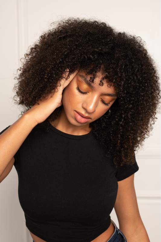 Woman with curly hair touching her temple and closing her eyes, appearing stressed in a neutral indoor portrait