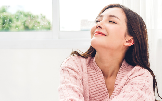 Young woman smiling with eyes closed near a bright window, appearing relaxed and content