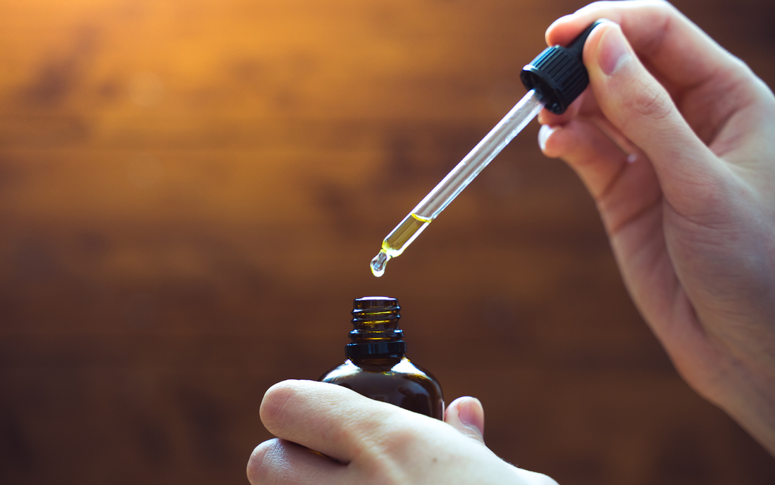 Hands holding a dropper above an amber oil bottle with a drop of yellow oil falling against a warm wooden background
