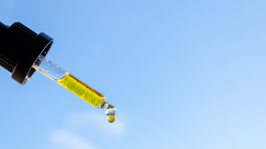 Close-up of a dropper holding golden THC oil with a droplet forming against a clear blue sky background