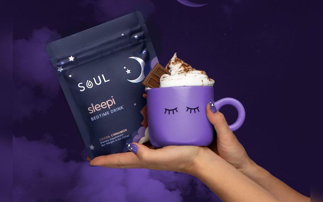 Front view of Soul Sleepi bedtime drink pouch and purple mug with whipped cocoa held in hands on purple cloud background