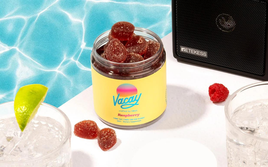 Jar of Vacay raspberry gummies on a white table by a pool backdrop with a lime-topped drink and a speaker