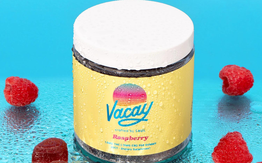 Close-up of Vacay Raspberry gummy jar with yellow label, 1.5mg THC and 15mg CBD, on wet blue background with raspberries