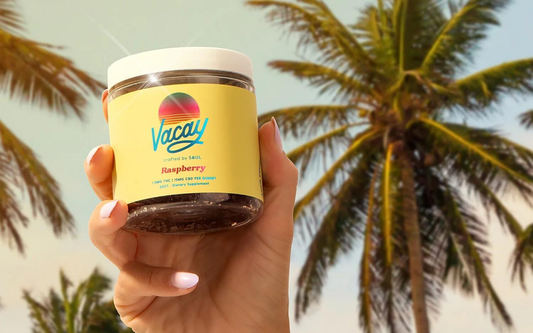 Close-up front view of Vacay Raspberry gummy jar showcasing yellow label and logo held in a hand against palm trees background