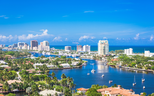 Aerial view of a sunlit Florida waterfront with canals, sailboats, and high-rise condos under a blue sky