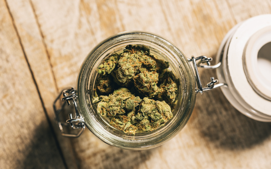 Top-down view of dried hemp flower buds in a glass jar showing dense green and orange trichomes on a wooden table