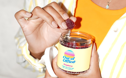 Close-up of an Out of Office raspberry gummies jar with a hand picking a sugar-coated raspberry gummy against an orange top