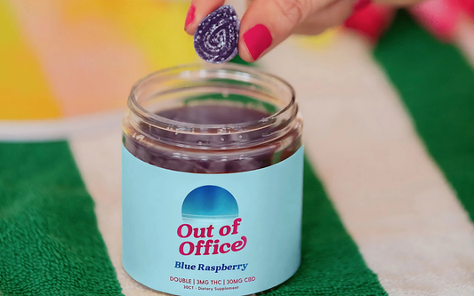 Close-up of Out of Office Blue Raspberry gummy jar showing the open jar and a purple gummy held above it on a green-and-white towel