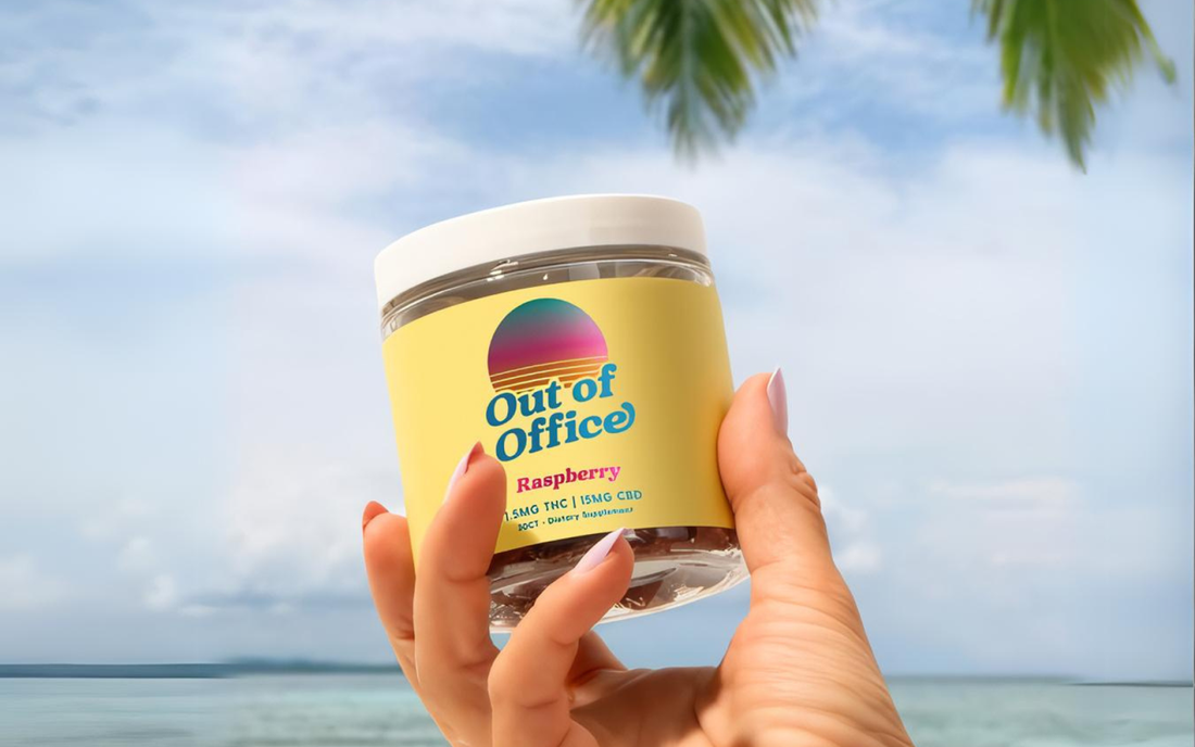 Angled view of Out of Office Raspberry jar showcasing yellow label with retro sunset logo, clear glass jar and white lid held against a beach background